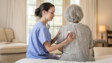 A healthcare professional and another person sitting on a bed with the healthcare provider listening to the other's lungs using a stethoscope