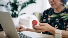 A woman holding a pill bottle and using a laptop.