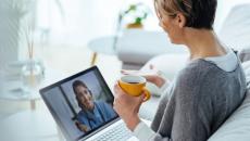 Person sitting on their couch holding a cup of coffee while speaking to a virtual healthcare provider on their computer