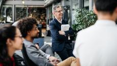 A person standing up, holding paperwork watching something while two other people are sitting and one other person has their back turned 