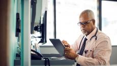 "Healthcare professional sitting at a desk with a window behind them while wearing a stethoscope around their neck and looking at a tablet"