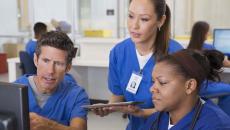 Three healthcare providers wearing scrubs sitting around a computer looking at the screen