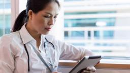 Healthcare provider sitting near a window while wearing a stethoscope and looking at a tablet