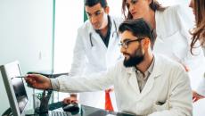 Three people wearing lab coats with one seated and pointing at screen