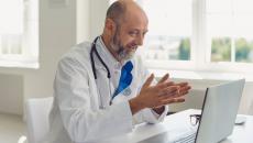 Healthcare provider sitting at a desk wearing a lab coat and a stethoscope while speaking to someone on a computer