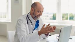 Healthcare provider sitting at a desk while wearing a lab coat and a stethoscope around their neck and speaking with someone on a computer