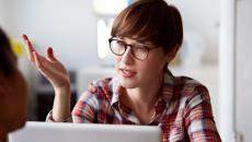 A person with a red plaid shirt on with short hair wearing glass, sitting at a computer with their hand up while talking